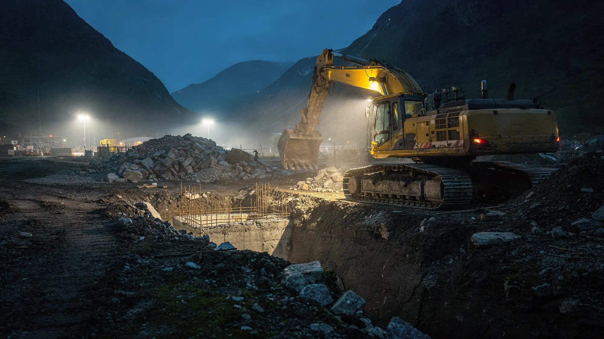 En gravemaskin graver ut en byggegrop på en anleggsplass i skumringen, med arbeidslys og fjell i bakgrunnen.